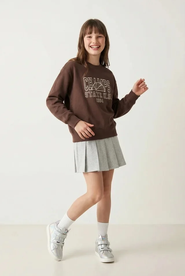 A child fashion model wearing a brown sweatshirt, grey skirt, and boots in a catalog photograph